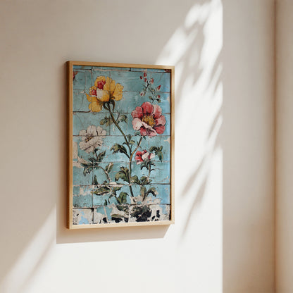 Framed wall art hanging on a beige wall, featuring a rustic floral design with yellow, pink, and white flowers on a distressed light blue background. The artwork is enclosed in a light wood frame and photographed in soft natural light with leafy shadows cast across the wall.
