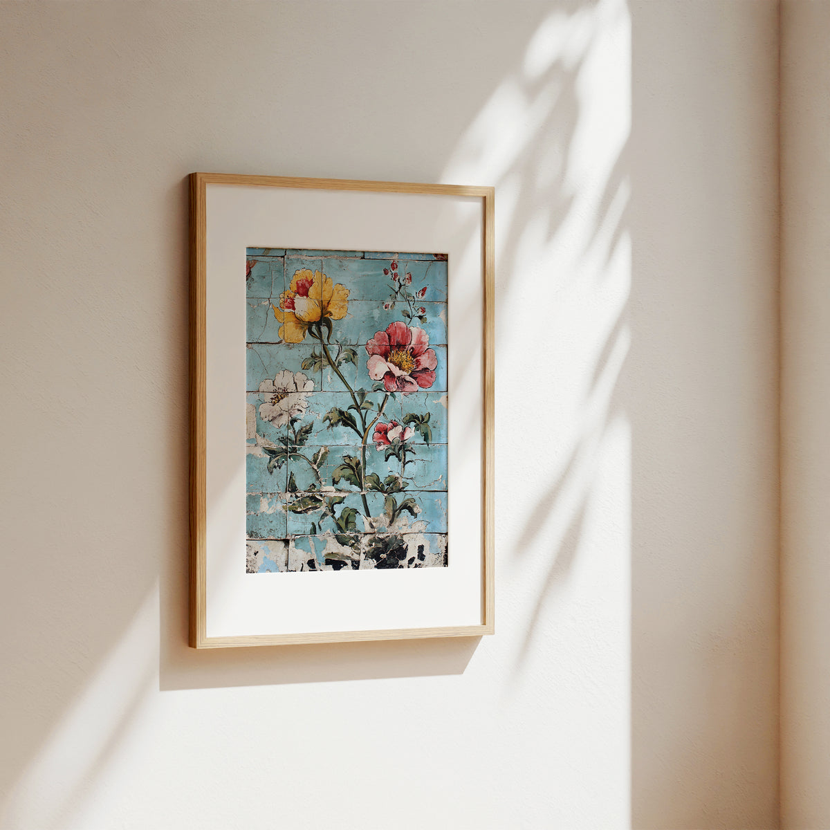 A framed floral art print with a light wood frame and white mat, hanging on a beige wall with soft sunlight and leaf shadows cast across it.
