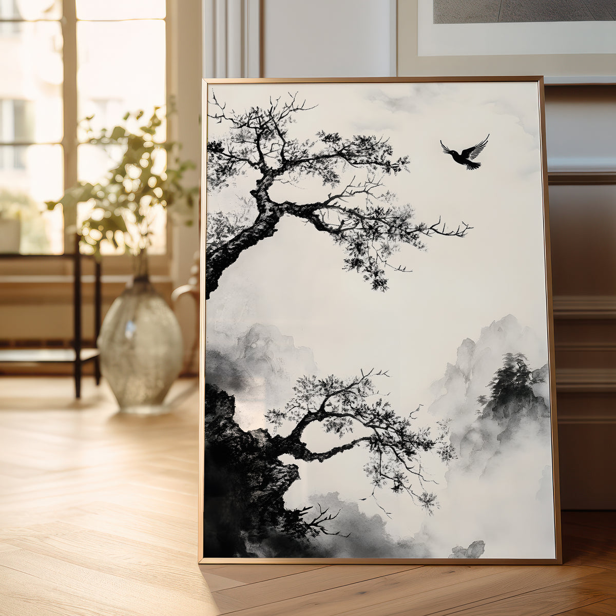 A vertical framed art print featuring a black and white Chinese ink-style landscape with gnarled trees, misty mountains, and a bird, displayed leaning against a wall on a light wood floor in a softly lit room.