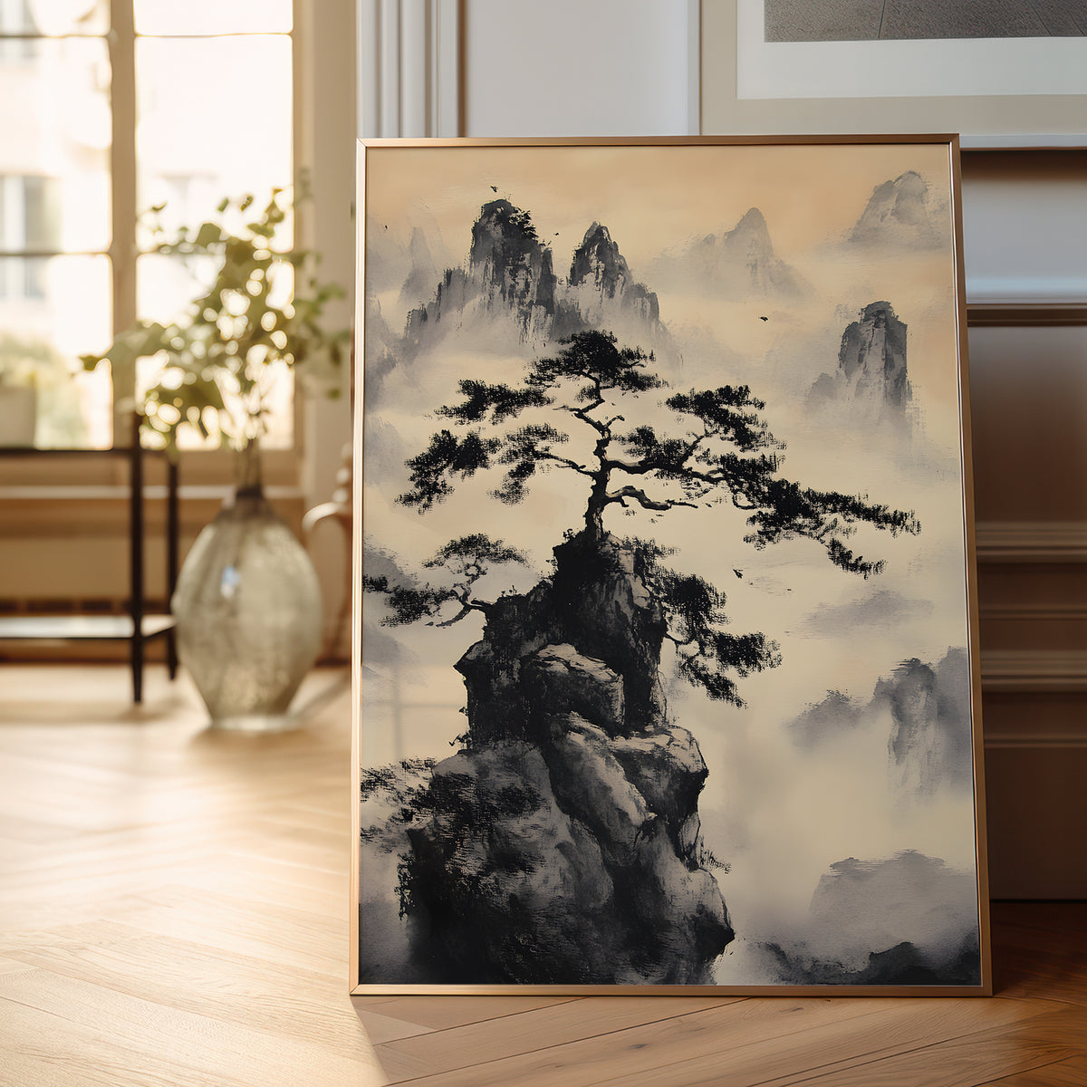 A framed vertical art print featuring a black ink-style pine tree on a rocky peak surrounded by misty mountains, leaning against a wall on a light wood floor in a softly lit interior.