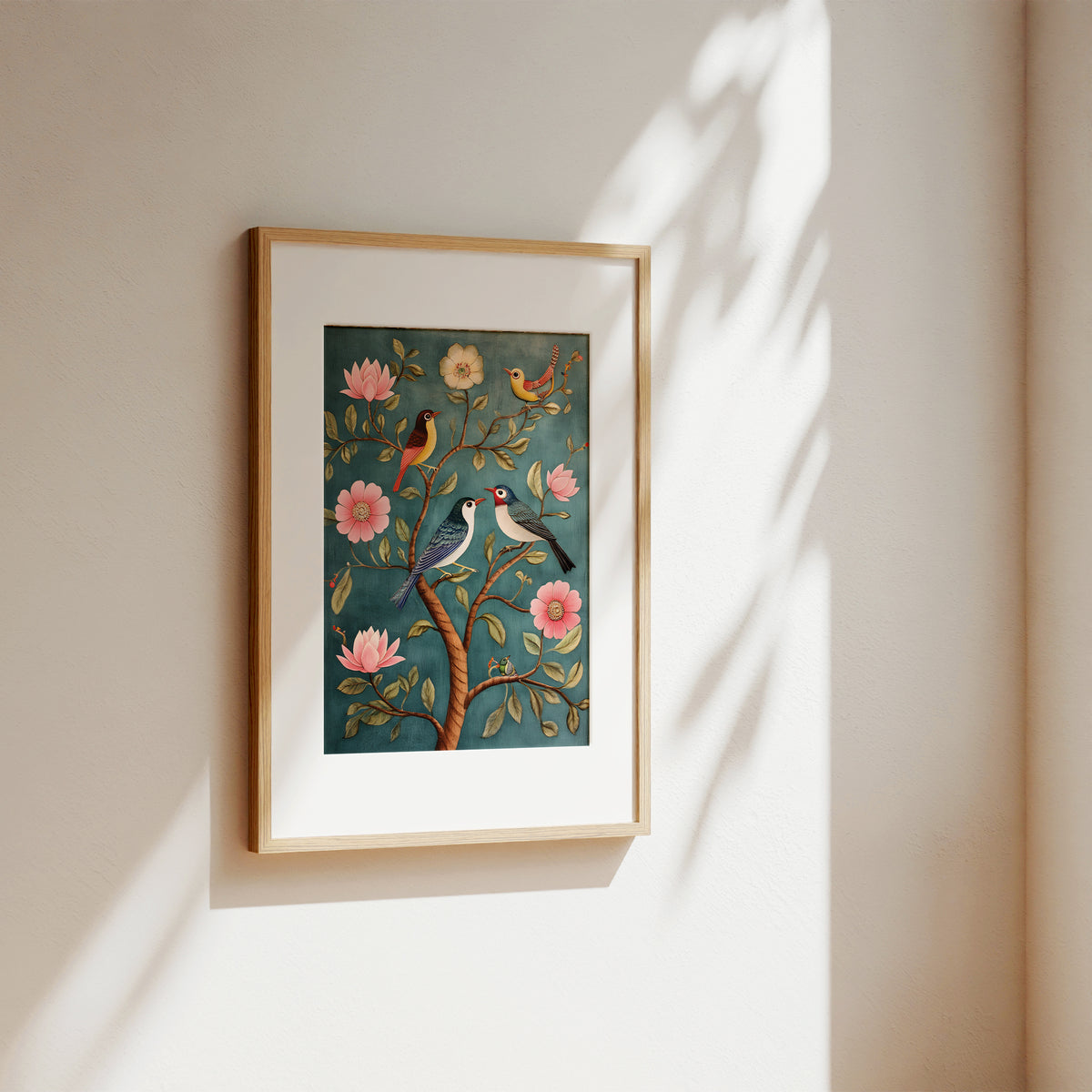 A framed art print featuring two birds perched on a tree with pink flowers, displayed on a warm beige wall with soft sunlight and leaf shadows.