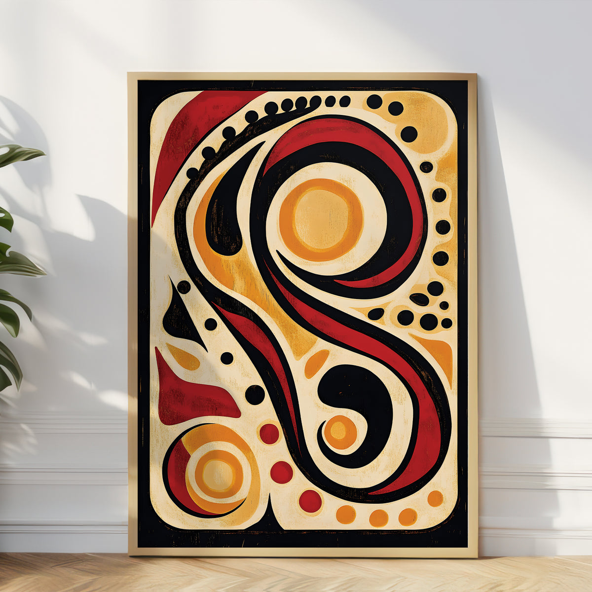 A framed abstract art print leaning against a white wall on a light wood floor, featuring bold organic swirls and circles in red, black, cream, and orange.