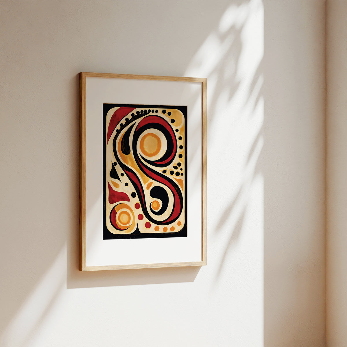 Framed abstract wall art hanging on a warm beige wall, with sunlight casting leaf shadows across the surface. The artwork features bold black, red, yellow, and cream curved shapes inside a black rectangle, surrounded by a white mat and light wood frame.