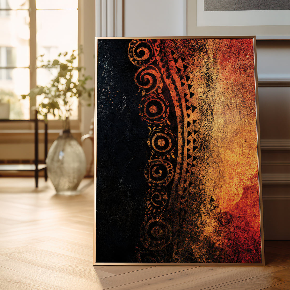 A framed vertical art print leaning against a wall on a light wood floor in a bright, modern room. The artwork features a textured design split diagonally, with a dark black-and-brown tribal pattern on the left and a warm orange, gold, and red abstract texture on the right.