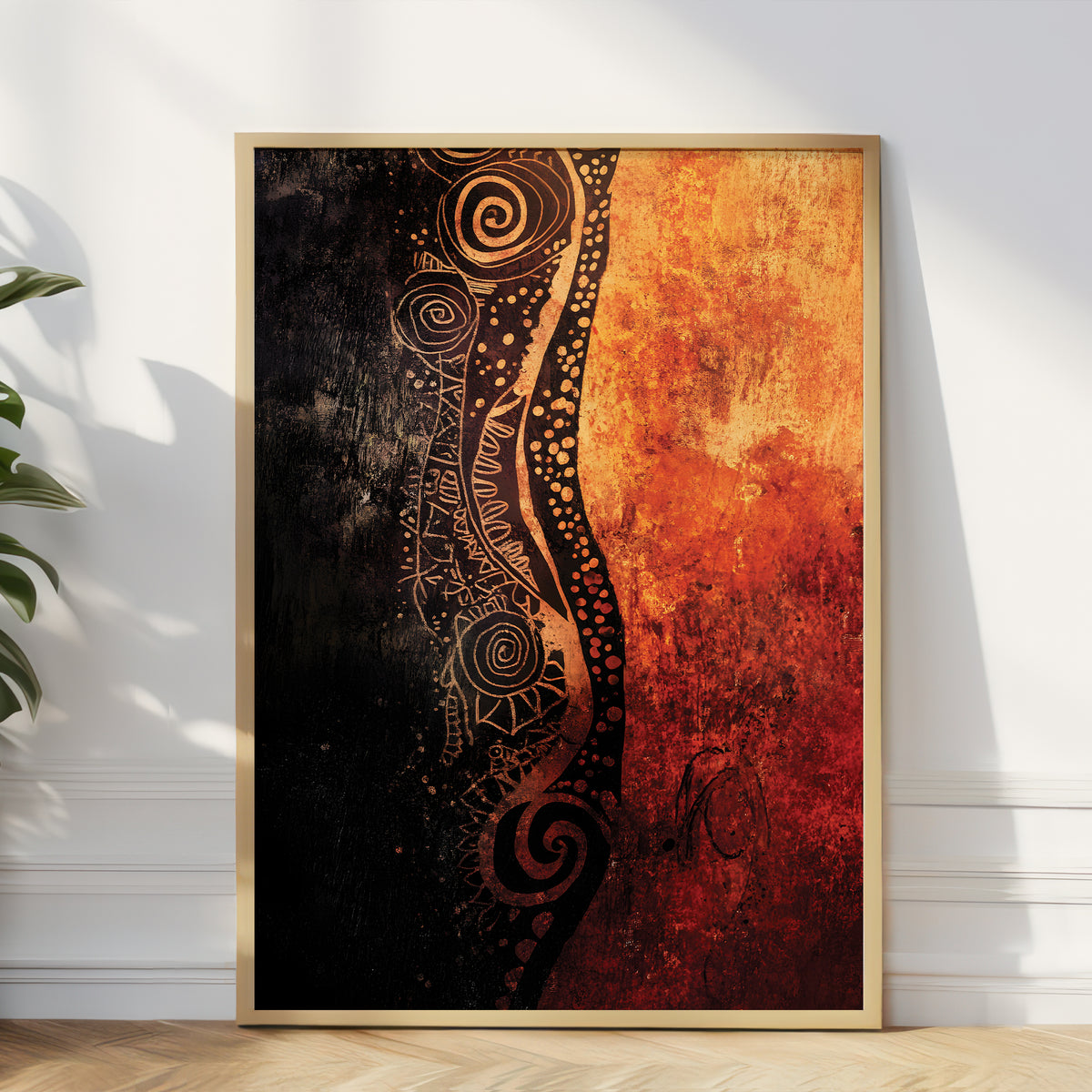 A framed abstract art print leaning against a white wall on a light wood floor, with a plant leaf visible at the left edge. The artwork is split vertically into a dark black-and-brown patterned left side and a warm orange-red textured right side, enclosed in a thin light wood frame.