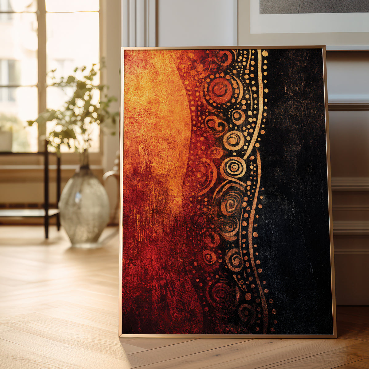 A framed abstract art print leaning against a wall on a light wood floor in a bright room. The artwork features a textured vertical composition with a warm orange and red left side, a black right side, and a decorative gold circular pattern separating the two sections.