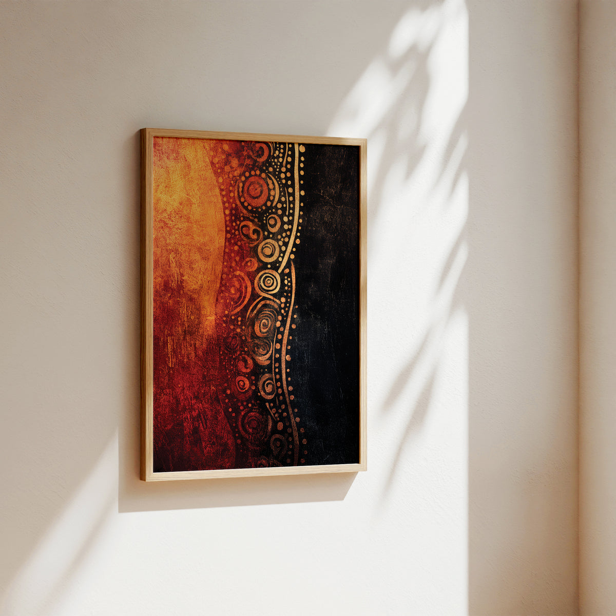 A framed abstract wall art print hanging on a beige wall, featuring a vertical composition with warm orange and red textures on the left and a black background with gold circular dot patterns on the right. The frame is a light natural wood, and the scene is lit by soft sunlight with leaf shadows cast on the wall.