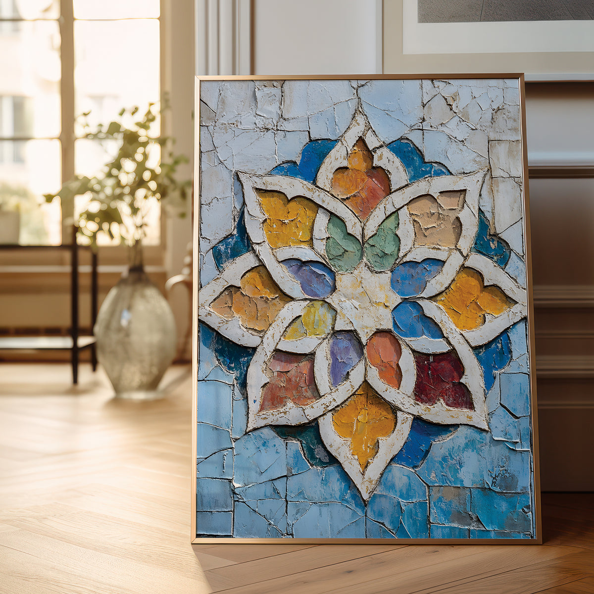 A framed square wall art piece leaning against a wall on a light wood floor in a bright interior. The artwork shows a colorful geometric floral medallion with cracked texture, blue background tiles, and white outlines, with warm yellow, orange, green, purple, and red sections.
