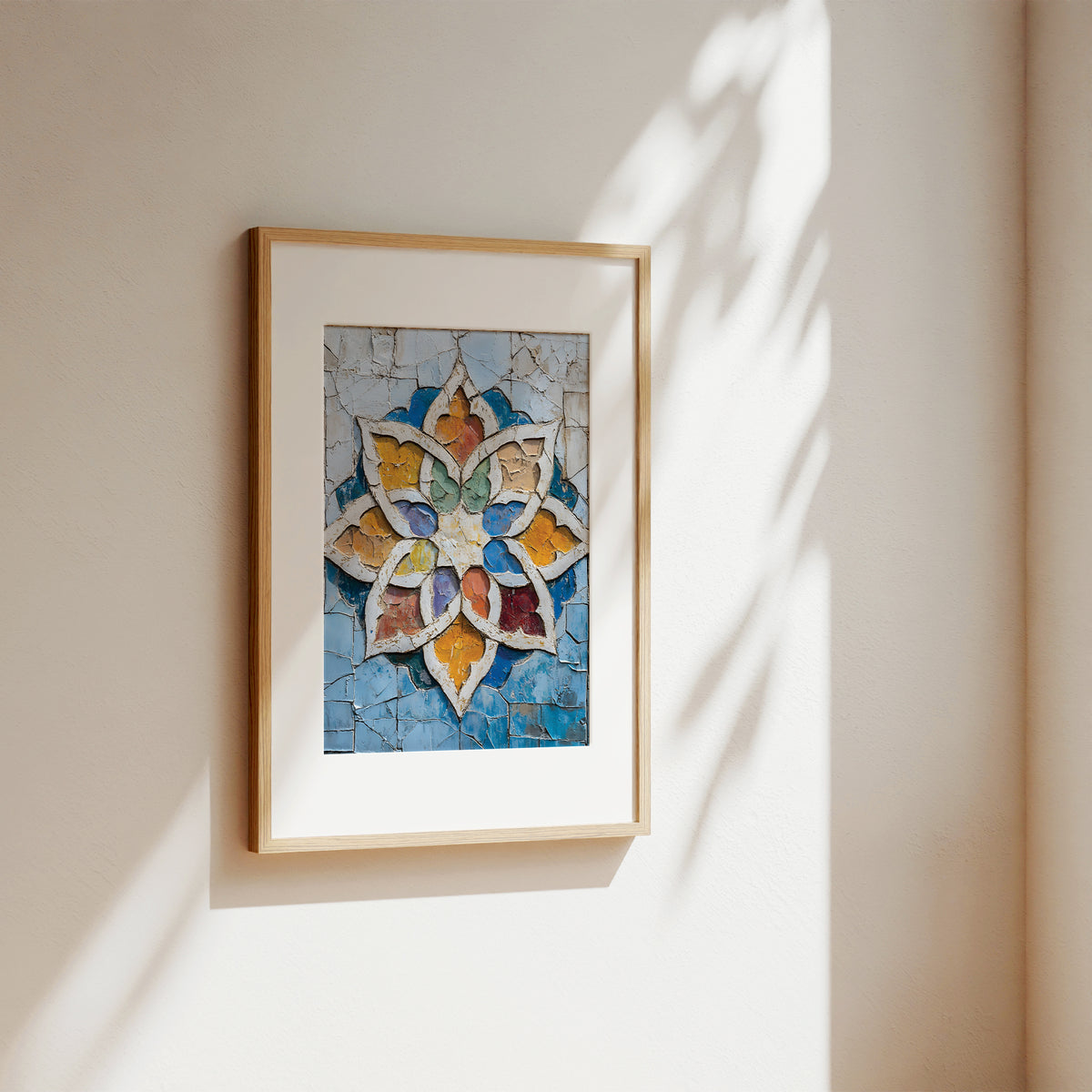 A framed art print featuring a colorful mosaic-style floral or starburst design in blue, gold, white, and red tones, hanging on a warm beige wall with soft sunlight and leaf shadows cast across it.