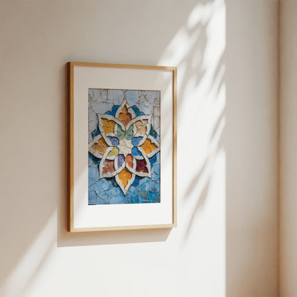 A framed art print featuring a colorful mosaic-style floral or starburst design in blue, gold, white, and red tones, hanging on a warm beige wall with soft sunlight and leaf shadows cast across it.