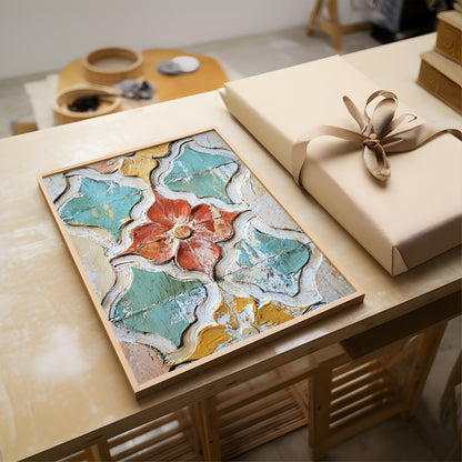 A framed abstract floral artwork with textured teal, cream, mustard, and coral tones sits on a light wood table beside a wrapped gift box with a beige ribbon.