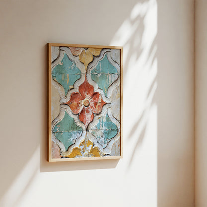 A framed decorative wall art piece hanging on a beige wall, featuring a distressed geometric floral pattern in muted teal, terracotta, cream, and ochre tones. Sunlight casts soft shadows across the wall and frame.
