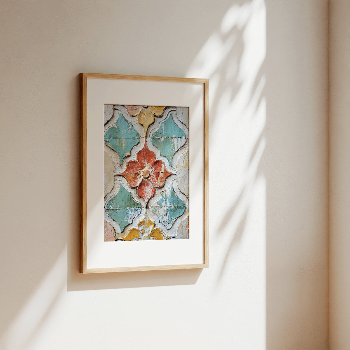 A framed art print featuring a colorful, distressed mosaic tile pattern with a central red-orange flower, displayed on a warm beige wall with soft sunlight and leaf shadows.