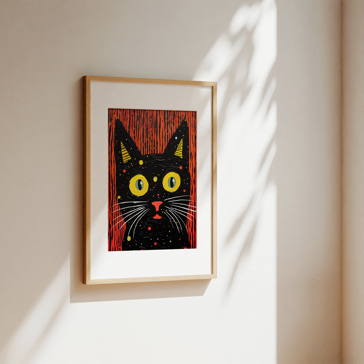 A framed art print featuring a stylized black cat with yellow eyes and a red background, hanging on a warm beige wall with soft sunlight and leaf shadows.