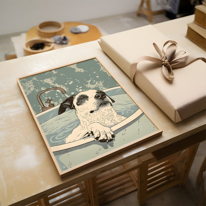 A framed art print featuring a dog relaxing in a bathtub, styled on a wooden table next to a wrapped gift box with a beige ribbon.
