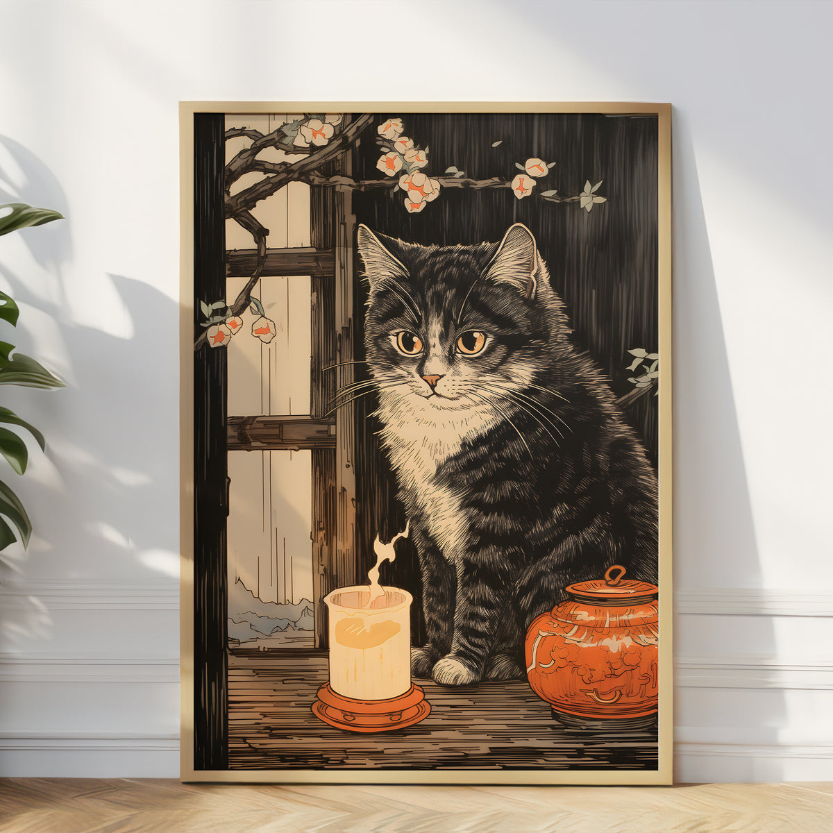 A framed vertical art print leaning against a white wall on a wood floor. The artwork shows a black-and-white tabby cat beside a glowing candle and an orange ceramic jar, with cherry blossoms and a window in the background.