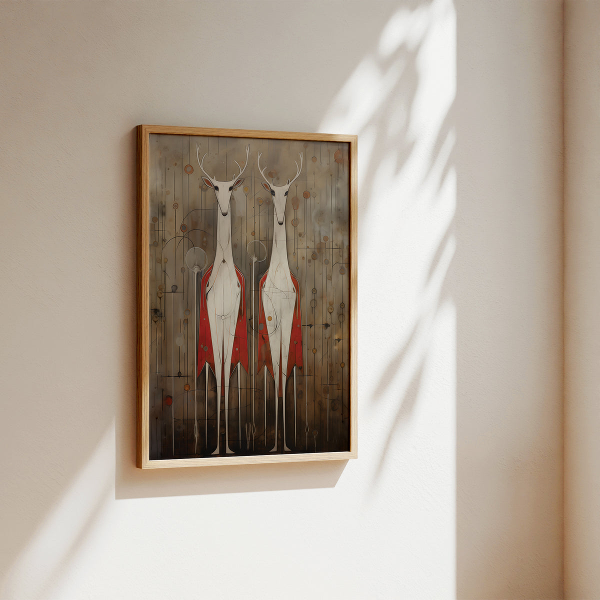A framed art print featuring two stylized white deer with red garments standing against a rustic wood background, hanging on a beige wall with soft sunlight and leaf shadows.