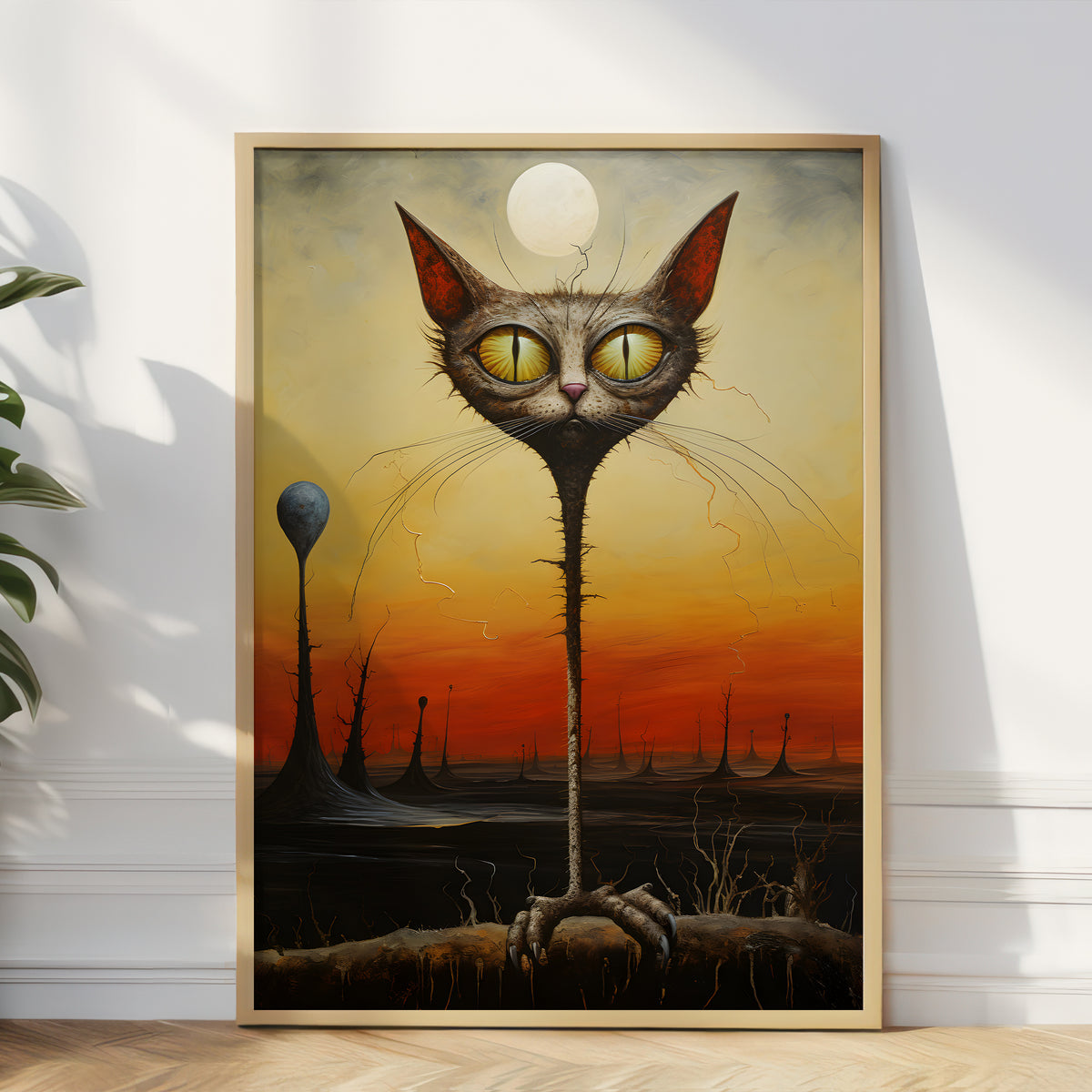 A framed surreal art print leaning against a white wall on a light wood floor, showing a tall gray cat with large yellow eyes and a long neck standing in a dark landscape under a glowing orange sky.