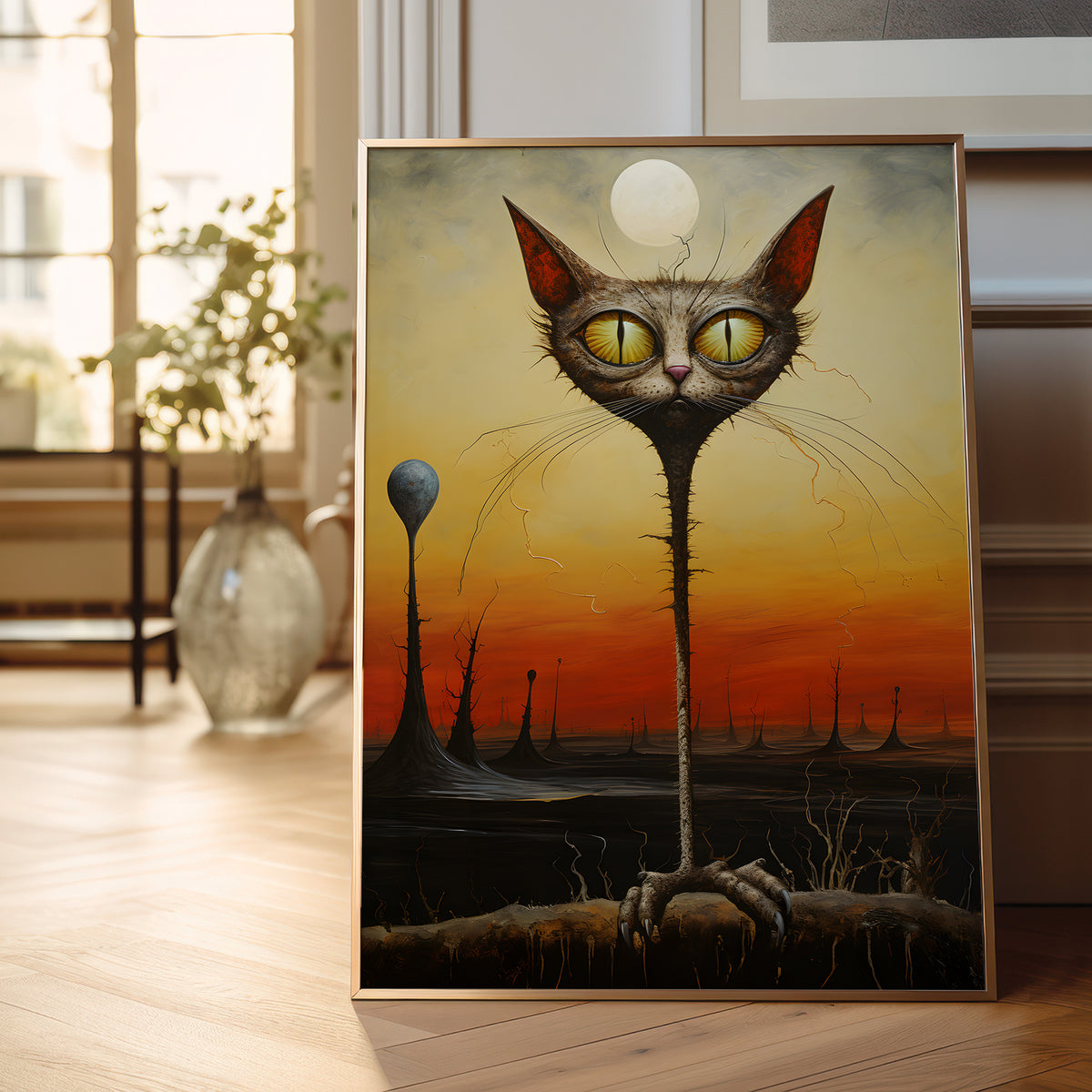 A framed surreal art print leaning against a wall on a wooden floor in a bright room. The artwork shows a large cat head with glowing yellow eyes emerging from a dark landscape under a sunset sky, with a pale moon and tall spire-like forms in the background.