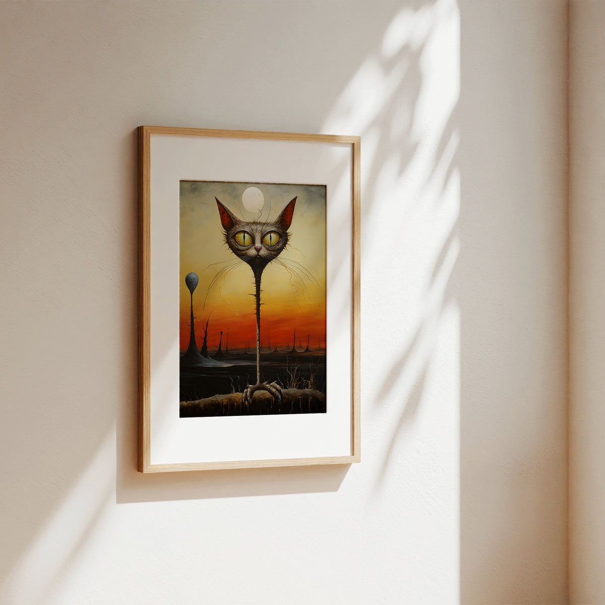 A framed surreal art print hanging on a warm beige wall, featuring a tall spindly cat head with large yellow eyes over a dark landscape at sunset. The print has a white mat and a light natural wood frame, with soft shadow patterns cast across the wall.