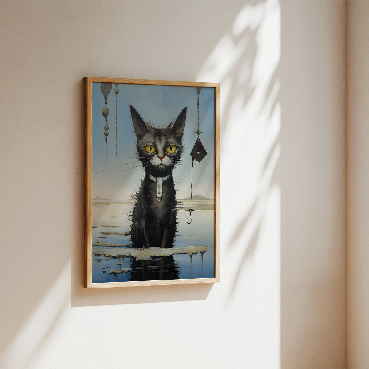 A framed surreal art print of a black cat with yellow eyes sitting in shallow water, hanging from a hook with a keychain, displayed on a warm beige wall with soft sunlight and leaf shadows.