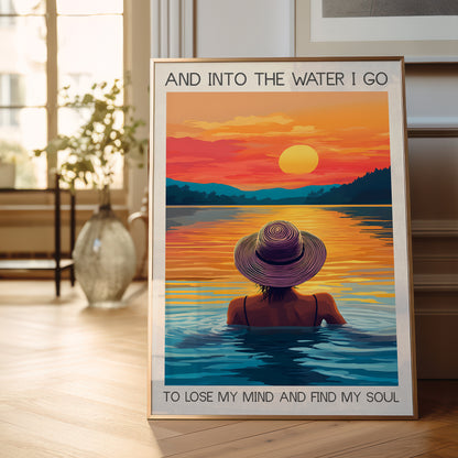 Framed poster of a person in a hat sitting in water with a sunset over a lake, placed on a wooden floor.