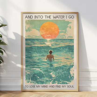 Framed artwork of a person in water with a sunset, text 'And into the water I go / To lose my mind and find my soul', on a white wall.