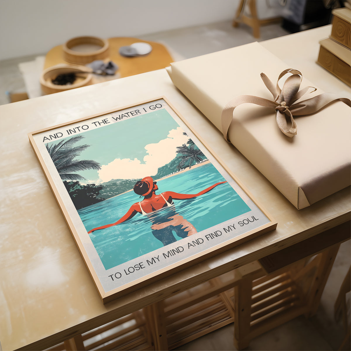 Framed artwork of a woman in a bikini floating in water with text, placed on a wooden surface next to a wrapped gift.