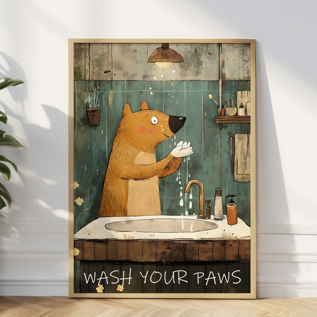 A framed vertical art print leaning against a white wall in a room. The print shows a cartoon brown bear washing its paws at a sink in a bathroom, with the text 'WASH YOUR PAWS' at the bottom.