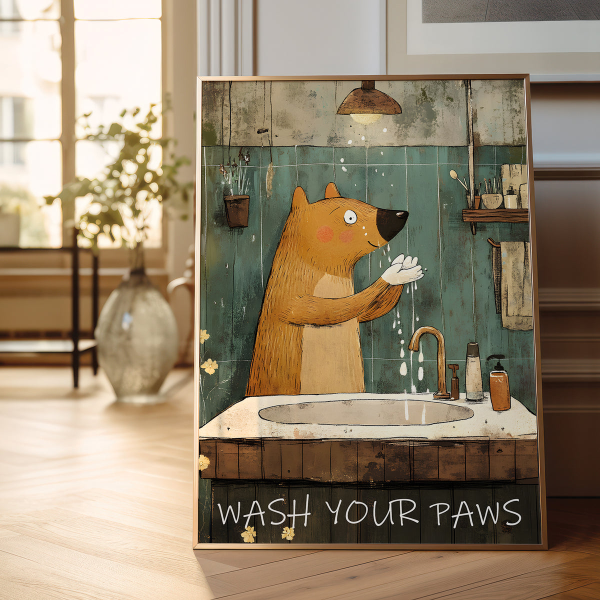 A framed art print featuring a cartoon brown bear washing its paws in a rustic bathroom, with the text 'WASH YOUR PAWS' at the bottom, displayed on a wooden floor in a bright room.