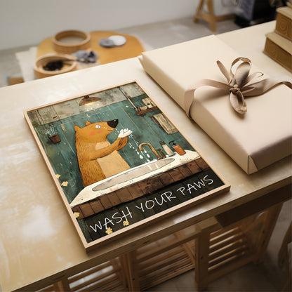 A framed art print featuring a cartoon brown bear washing its paws at a sink, with the text 'WASH YOUR PAMS' at the bottom, displayed on a wooden table next to a wrapped gift box.