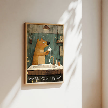 A framed art print featuring a cartoon brown bear washing its paws at a sink, hanging on a beige wall with soft sunlight and leaf shadows cast across it.