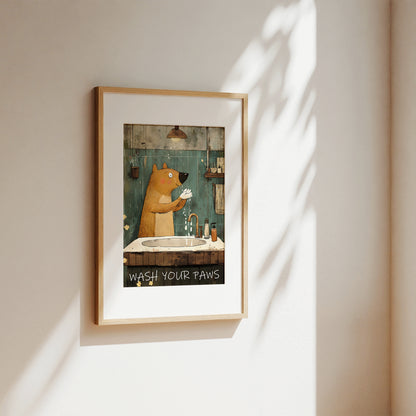 A framed art print featuring a cartoon brown bear washing its paws in a bathroom, hanging on a beige wall with soft sunlight and leaf shadows cast across it.