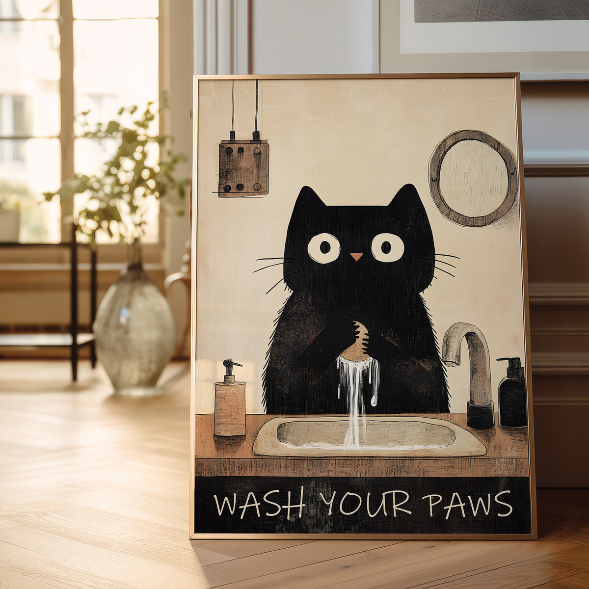 A framed art print featuring a whimsical black cat washing its paws at a sink, displayed on a wooden floor in a bright, modern room.