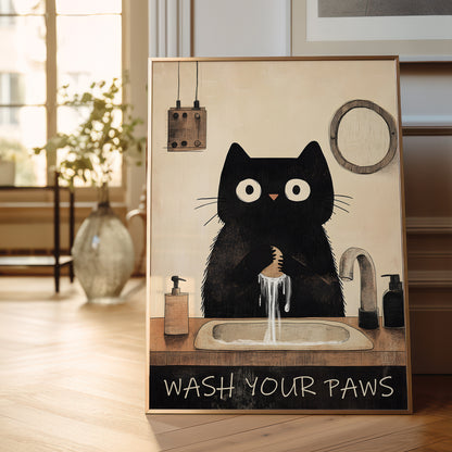 A framed art print featuring a whimsical black cat washing its paws at a sink, displayed on a wooden floor in a bright, modern room.