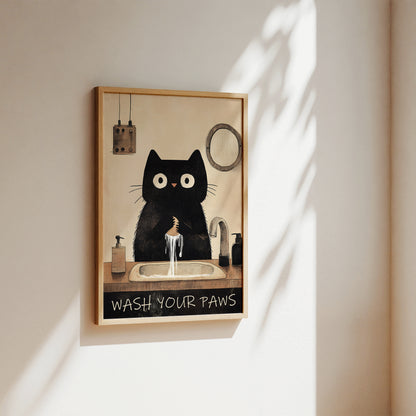A framed wall art print hanging on a beige wall, featuring a black cat sitting at a sink with running water and the text 'WASH YOUR PAWS'. The print has a light wood frame and is photographed in warm natural light with soft shadows cast across the wall.