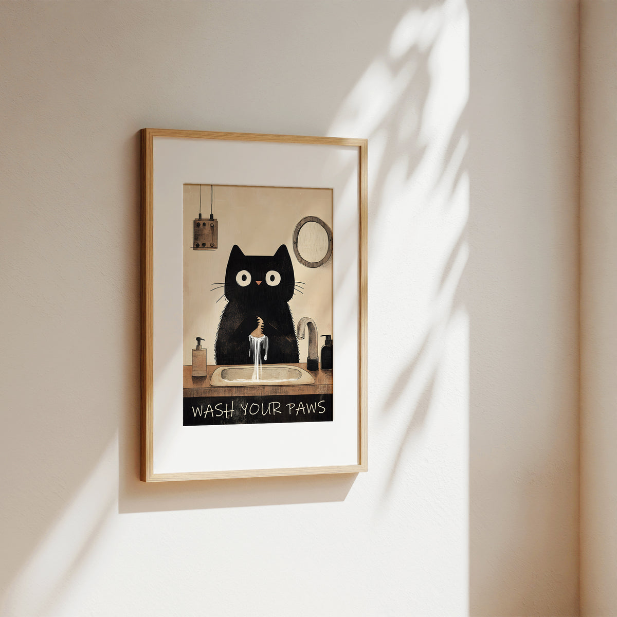 A framed art print featuring a black cat with large white eyes washing its paws at a sink, with the text 'WASH YOUR Paws' at the bottom. The print is housed in a light wood frame and displayed on a warm beige wall with soft, dappled sunlight casting shadows.