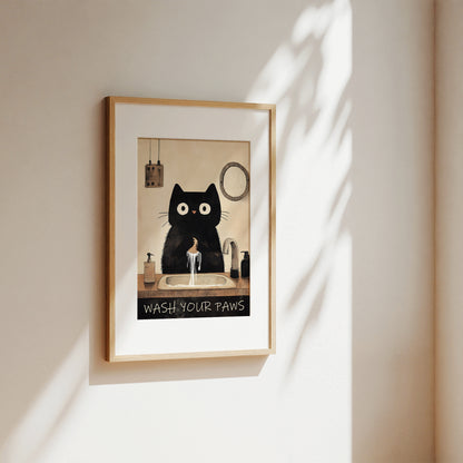 A framed art print featuring a black cat with large white eyes washing its paws at a sink, with the text 'WASH YOUR Paws' at the bottom. The print is housed in a light wood frame and displayed on a warm beige wall with soft, dappled sunlight casting shadows.