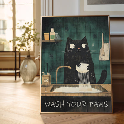 A framed art print leaning against a wooden wall in a bright room. The print shows a black cat washing its paws in a sink with the text 'WASH YOUR PAWS' at the bottom.