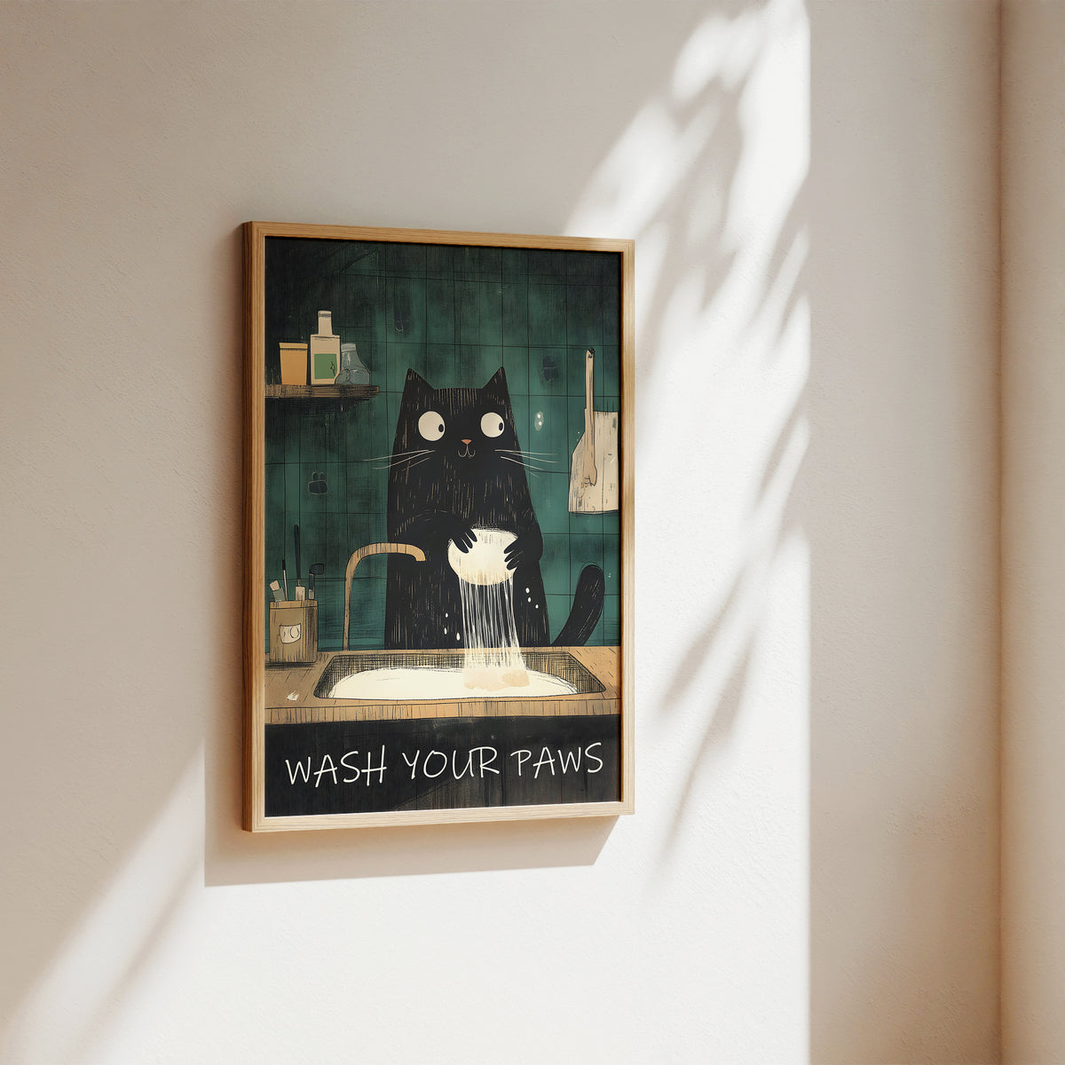 A framed wall art print hanging on a beige wall, showing a black cat washing its paws in a sink against a dark teal background. The print has a light wood frame and is photographed in warm natural light with soft shadows cast across the wall.