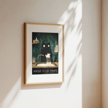 A framed art print featuring a black cat washing its paws at a sink, hung on a beige wall with soft sunlight and leaf shadows cast across it.