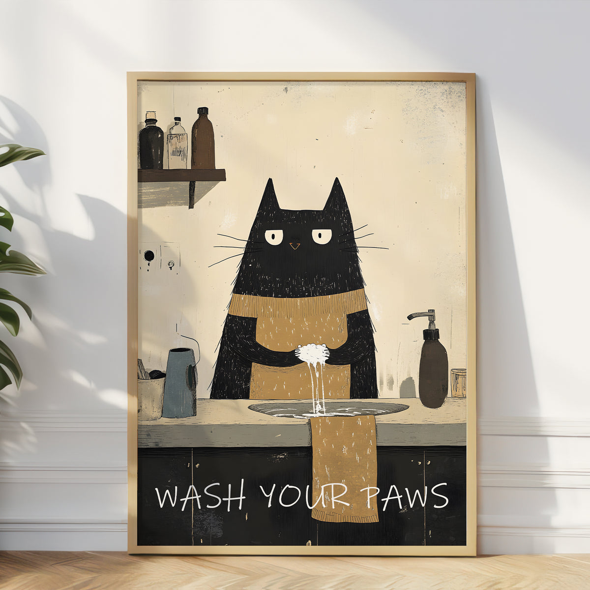 A framed art print leaning against a white wall on a light wood floor. The print shows a black cat with white eyes washing its paws at a sink, with the text 'WASH YOUR PAWS' at the bottom.