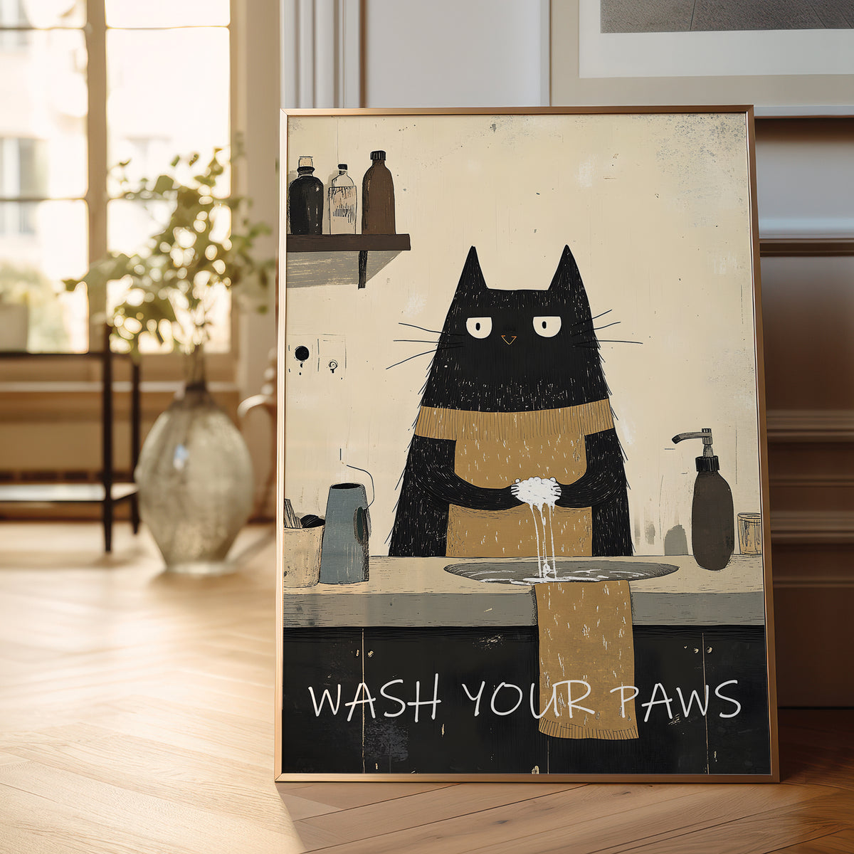 A framed art print featuring a black cat with white eyes washing its paws at a sink, with the text 'WASH YOUR PAWS' at the bottom. The print is displayed leaning against a wall on a light wooden floor in a bright, modern room.