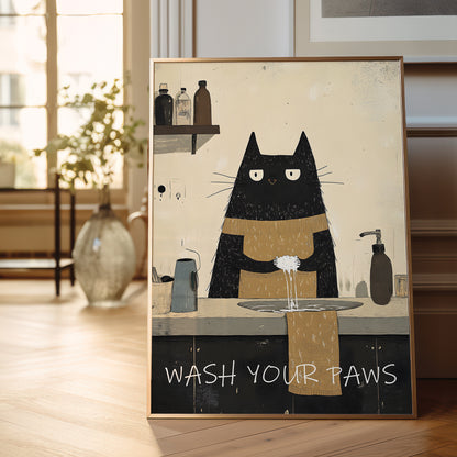 A framed art print featuring a black cat with white eyes washing its paws at a sink, with the text 'WASH YOUR PAWS' at the bottom. The print is displayed leaning against a wall on a light wooden floor in a bright, modern room.