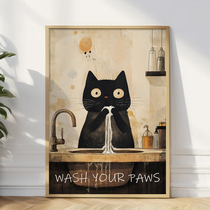 A framed wall art print leaning against a white wall on a wood floor. The artwork shows a black cat sitting at a sink with water coming out, with the text 'WASH YOUR PAWS' at the bottom.