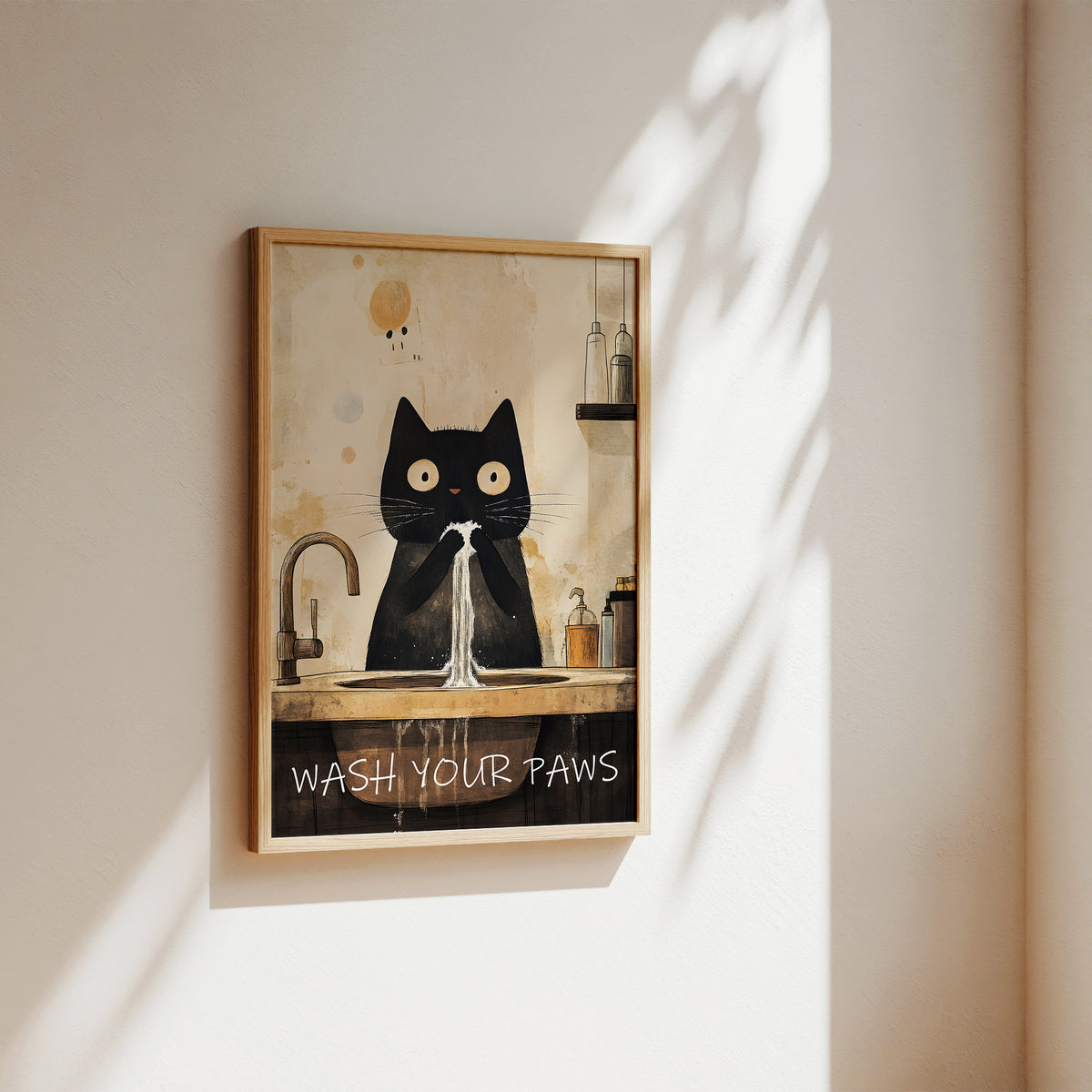 A framed wall art print hanging on a beige wall, showing a black cat sitting at a sink with the text 'WASH YOUR PAWS' below. The print has a warm cream background with a black cat illustration and a light wood frame.