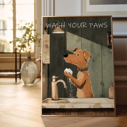 A framed art print leaning against a wall on a wooden floor in a bright room. The print shows a cartoon dog washing its paws at a sink, with the text 'WASH YOUR PAWS' at the top.