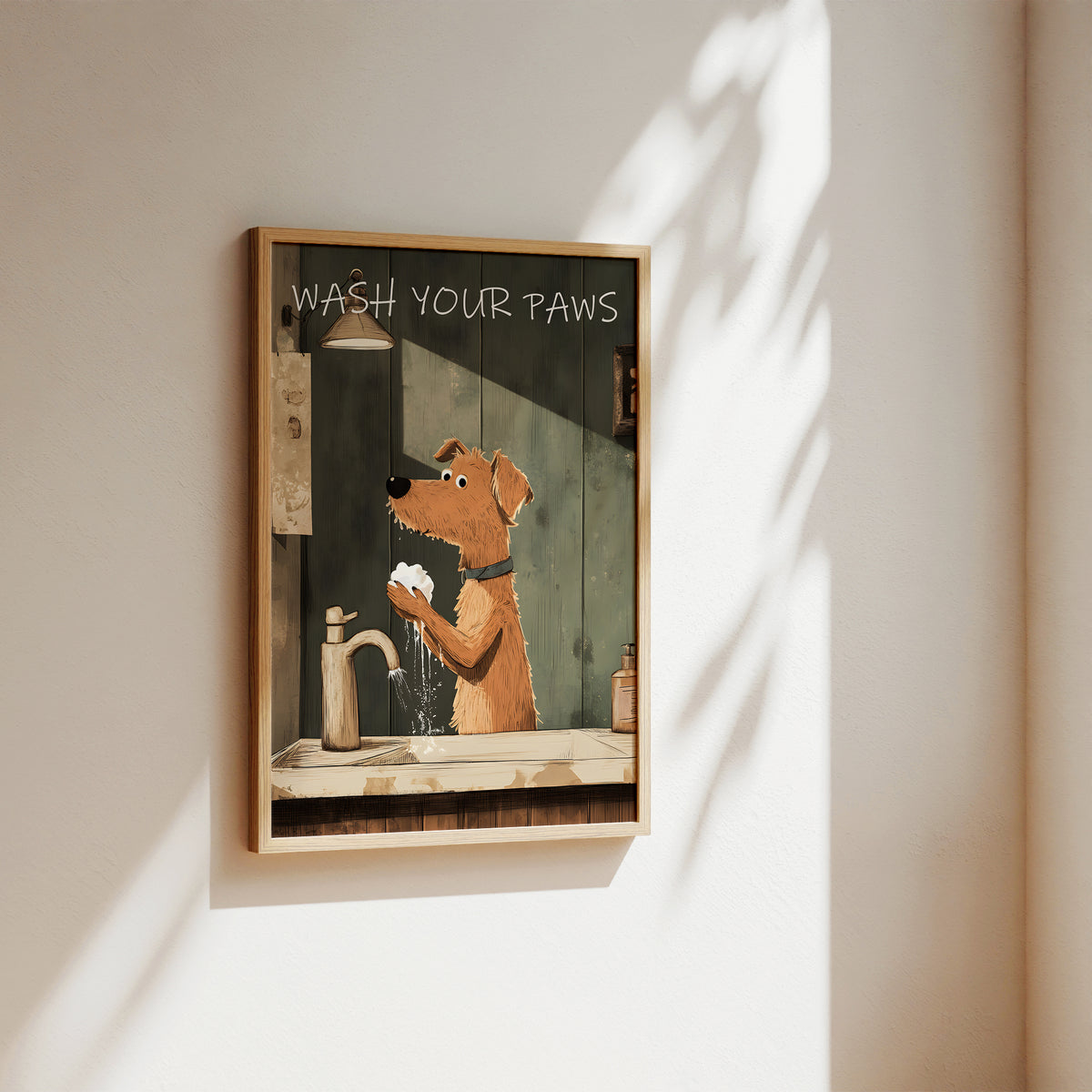 A framed wall art print hanging on a beige wall, showing an illustrated dog washing its paws with the text 'WASH YOUR Paws' at the top. The print is enclosed in a light wood frame and photographed in a real interior setting with sunlight and leaf shadows cast across the wall.