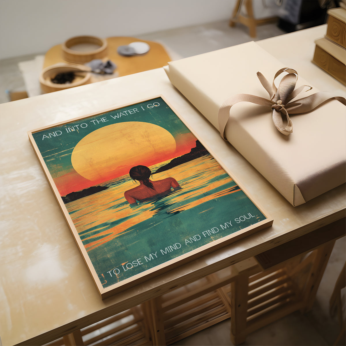 Poster of a person in water with a sunset and text, placed on a wooden surface next to a wrapped gift.