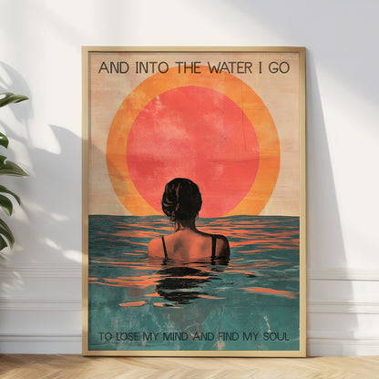 Framed artwork of a woman in water with a sunset, text 'And into the water I go / To lose my mind and find my soul' on a white wall.